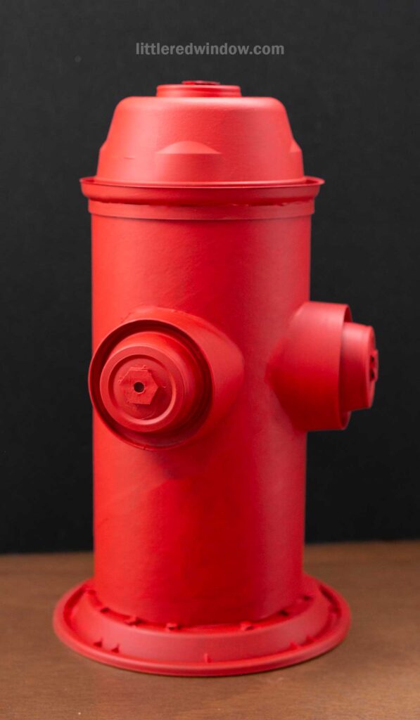 A small DIY cardboard fire hydrant, painted in bright red, stands on a brown surface against a black background.