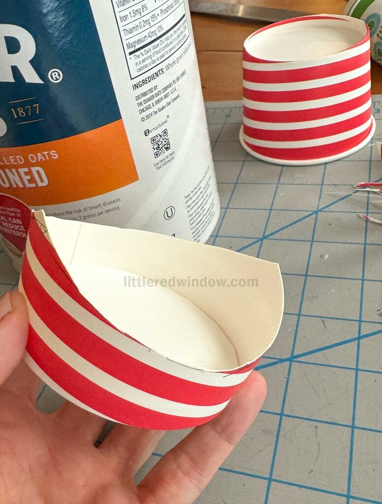 A hand holding a red and white striped paper cup, cut in half, with another cut cup in the background. An oatmeal container with visible text is nearby, all on a cutting mat surface.