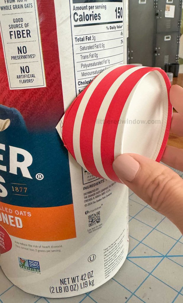 A person holds a red and white striped paper cup attached to the side of a large container of rolled oats, partially cut out to fit the side. The container's nutrition label is visible.