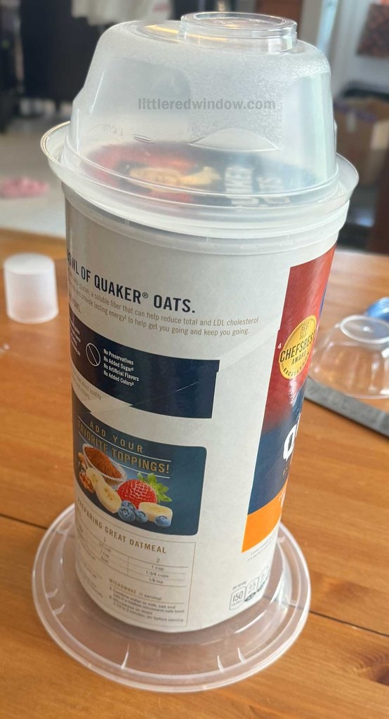 A large container of oats with a plastic lid and another plastic takeout lid as the base is placed on a wooden table.