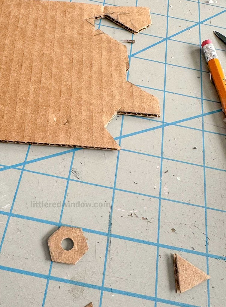 A cardboard piece with cut-out shapes, including a hexagon to look like a hardware store nut, lies on a gridded cutting mat. A sharpened pencil rests nearby.