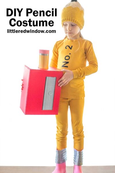 little girl in yellow pencil costume with pencil hat and pink boots holding a red cardboard box pencil sharpener with large pencil in it next to her waist