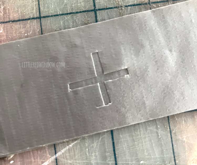a piece of silver duct tape with a plus shape cut out of it layered on top of another strip of silver duct take to look like a phillips head screw top