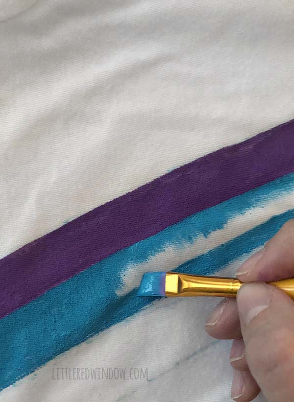 A hand painting a blue stripe of the rainbow on a white tshirt
