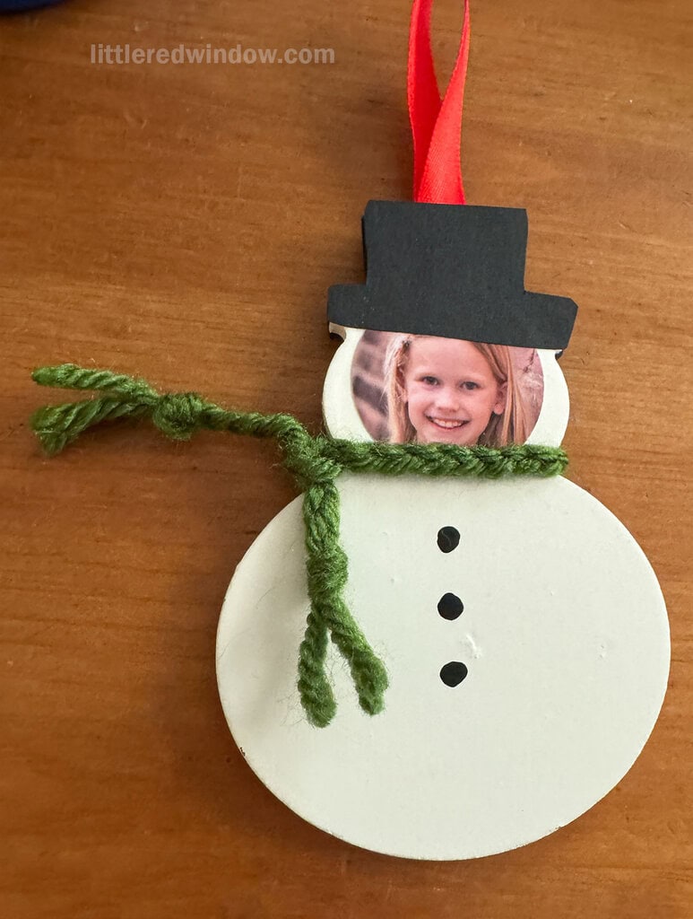 A handmade snowman ornament with a child’s photo as the face, a black paper top hat, green yarn scarf, three black buttons, and a red ribbon loop, lying on a wooden surface.