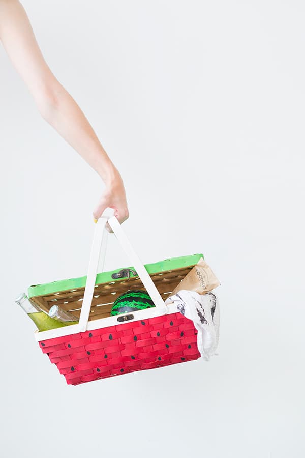 Womans arm holding wicker picnic basket with green painted lid and pink painted bottom with black seeds