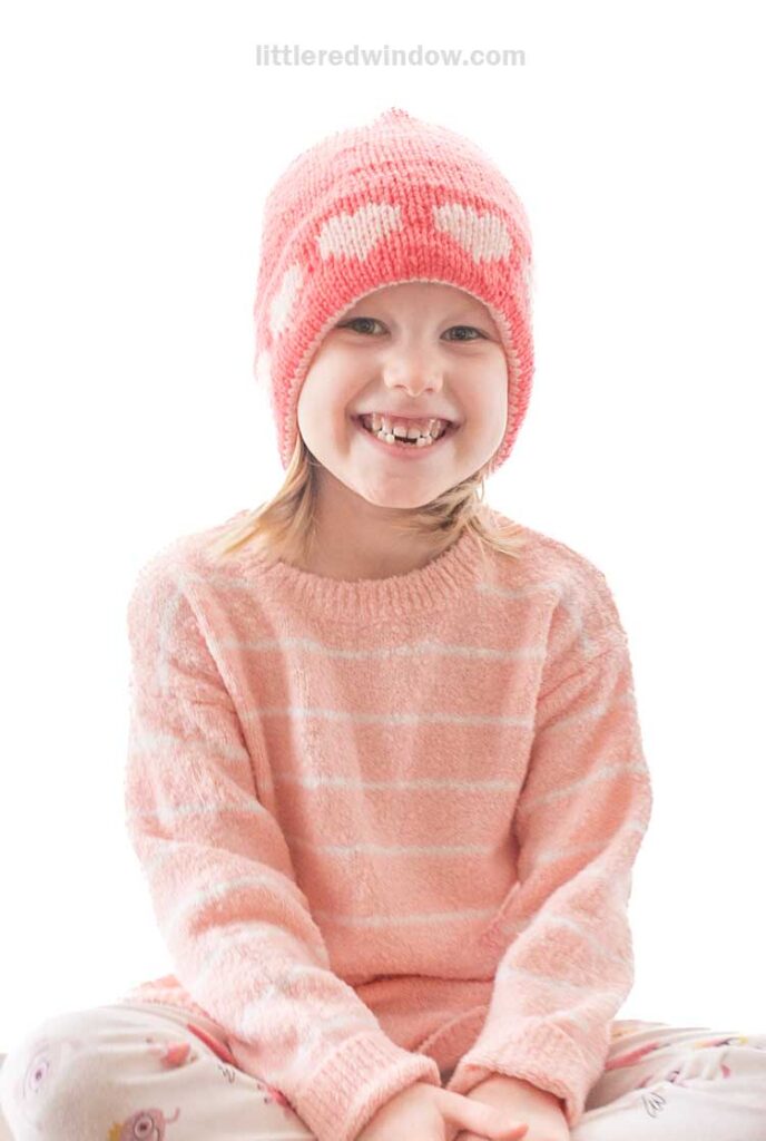 A smiling child is wearing a pink double brim heart hat paired with a matching pink striped sweater, sitting cross-legged. The bright and white background highlights their cheerful expression.