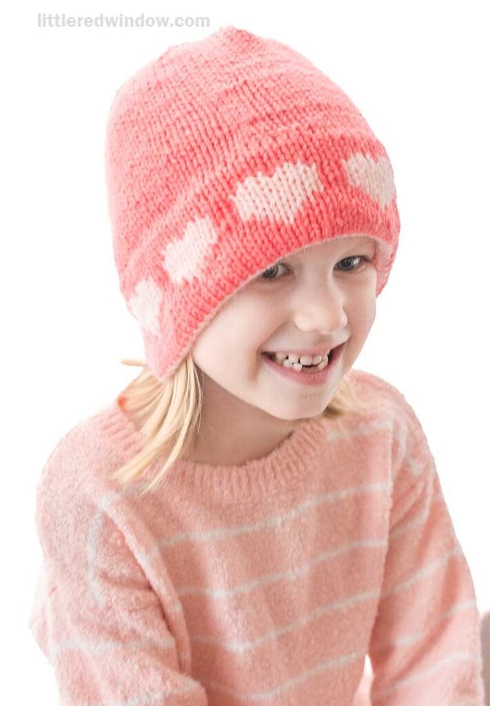 A smiling child wearing a pink double brim heart hat with white patterns and a matching fuzzy pink sweater with white stripes, set against a plain white background.