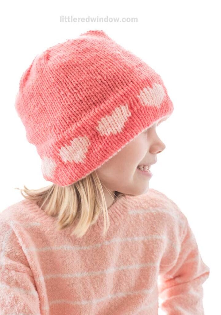 Child wearing a pink double brim heart hat with white hearts and a matching sweater, facing sideways and smiling against a white background.