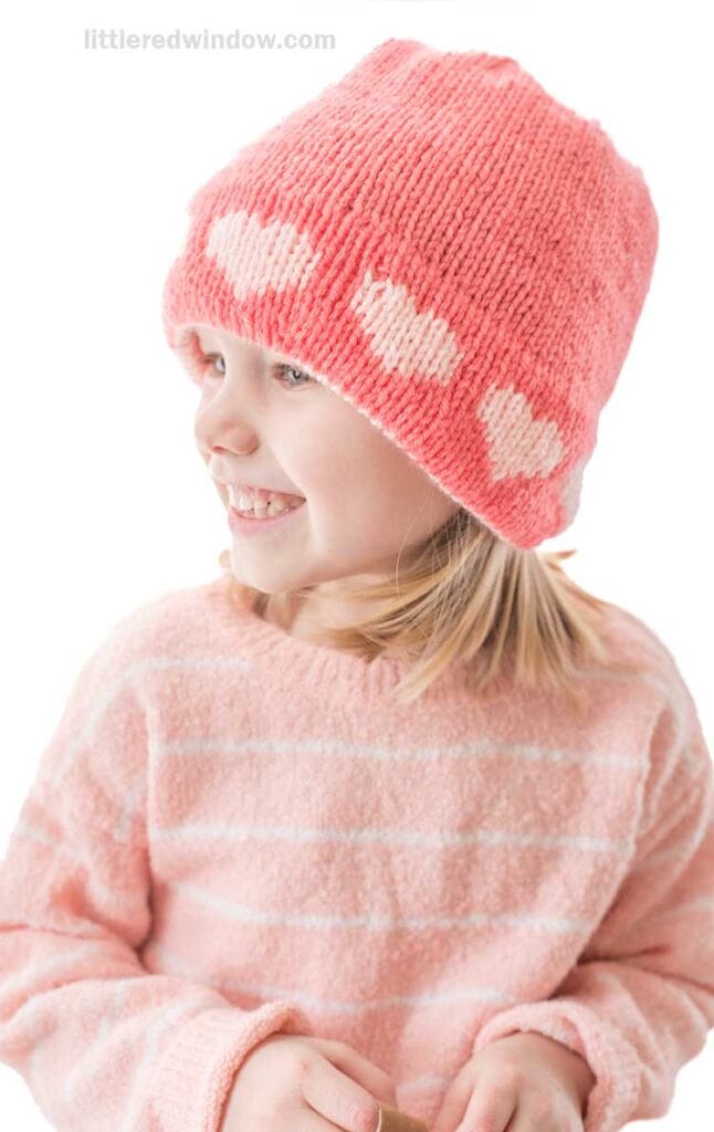 A young girl smiles warmly, sporting a pink double brim heart hat that complements her light brown hair. She's dressed in a cozy pink sweater against a plain white background.