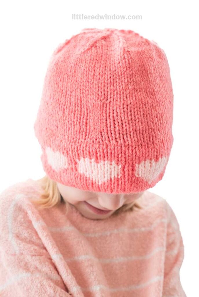 A child in a cozy pink knitted hat with double brim heart patterns glances downward. Complementing the look, they wear a charming pink and white striped sweater.