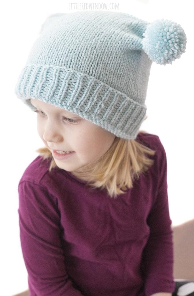 view from above of little blond girl in purple shirt wearing light blue knit hat with folded brim and two pom poms on top