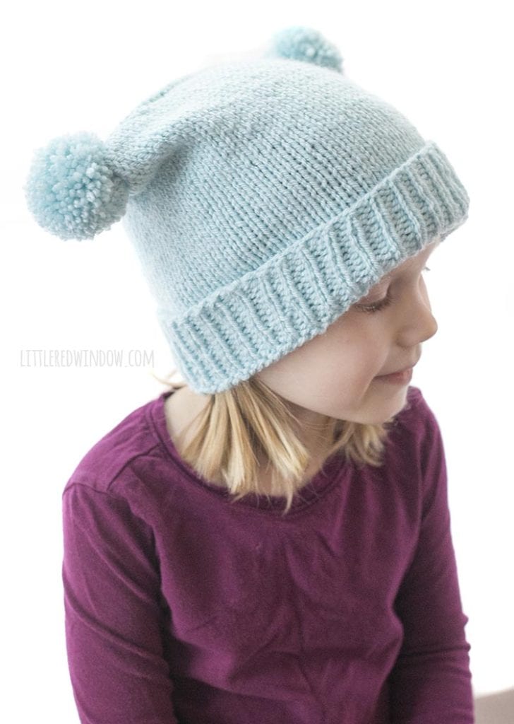 side view of double pom pom hat knitting pattern ona little girl in a purple shirt looking off to the right