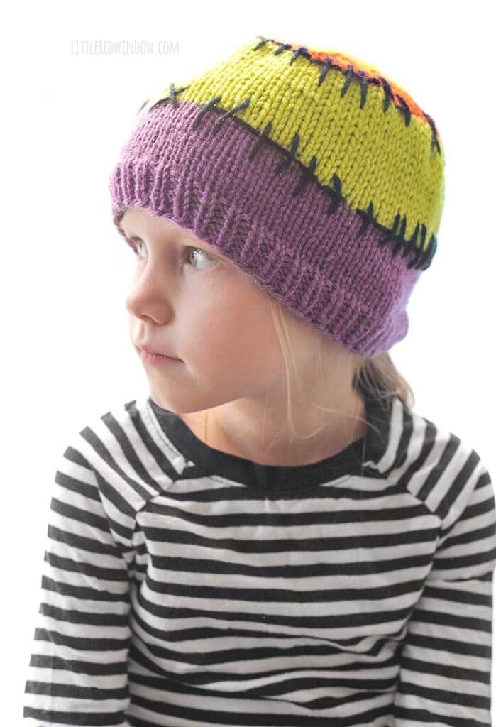 child in black and white striped shirt wearing a purple acid green and orange colorblocked knit hat with black stitching lines between the colors looking off in the distance to the left in front of a white background