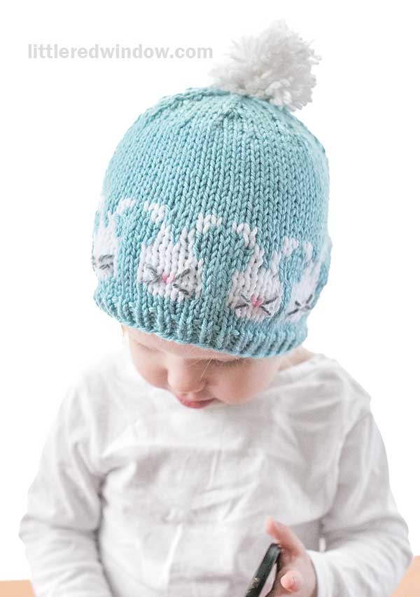 A young child is wearing an adorable Easter Bunnies hat—a knitted teal design with a white pom-pom on top and charming bunny patterns. The child, in a white shirt, gazes downward, capturing a moment of pure innocence.