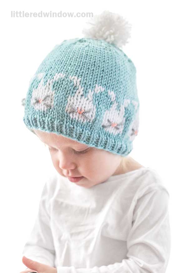 A small child wearing a light blue knit Easter Bunnies hat, adorned with whimsical white bunnies and a fluffy pom-pom on top. The child is dressed in a white shirt, gazing down with a neutral expression.