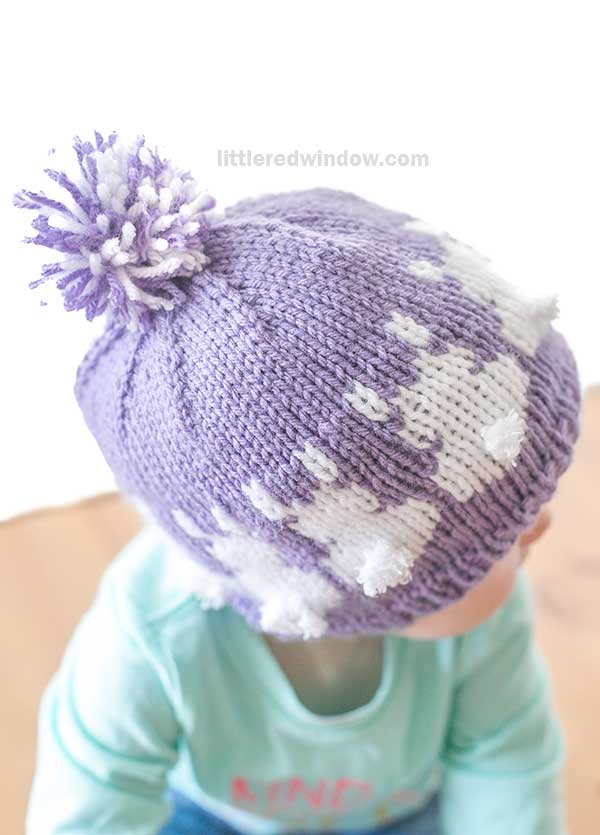 A baby sits on a soft brown surface, wearing a purple knit hat with white bunny patterns and a pom-pom, reminiscent of an Easter Bunny hat. Dressed in a light teal shirt, the little one is set against a white background with a faint website name above.