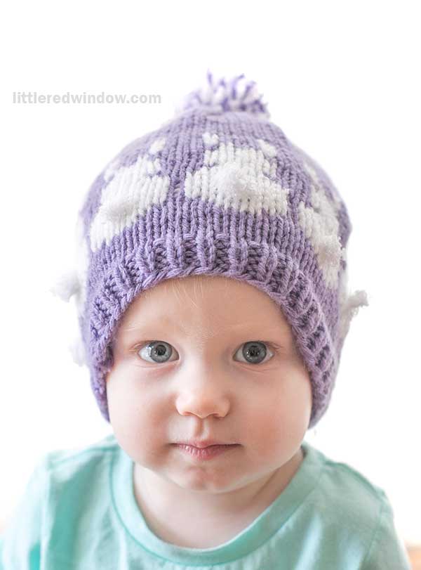 A baby wearing a purple knit Easter Bunny hat adorned with white bunny silhouettes and a pom-pom on top. The child, with light blue eyes and dressed in a mint green shirt, gazes directly at the camera with a neutral expression.