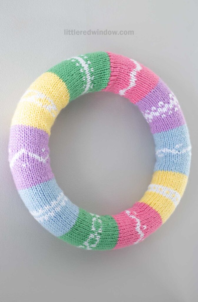 A knitted wreath with pastel striped sections in green, yellow, purple, pink, and blue with white zigzag patterns. The background is light gray.