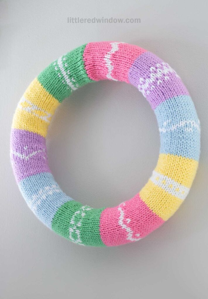 A colorful knitted wreath with sections in pastel pink, green, purple, blue, and yellow, each adorned with white geometric patterns, hanging on a light gray wall.