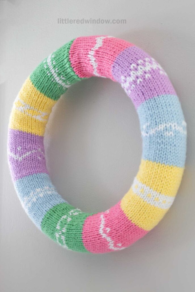 A knitted wreath with pastel stripes in pink, purple, yellow, green, and blue, featuring white patterned accents, hangs on a light gray wall.