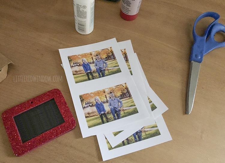 wood table with printed photos of two kids red glitter photo ornament scissor and two bottles of red and white craft paint