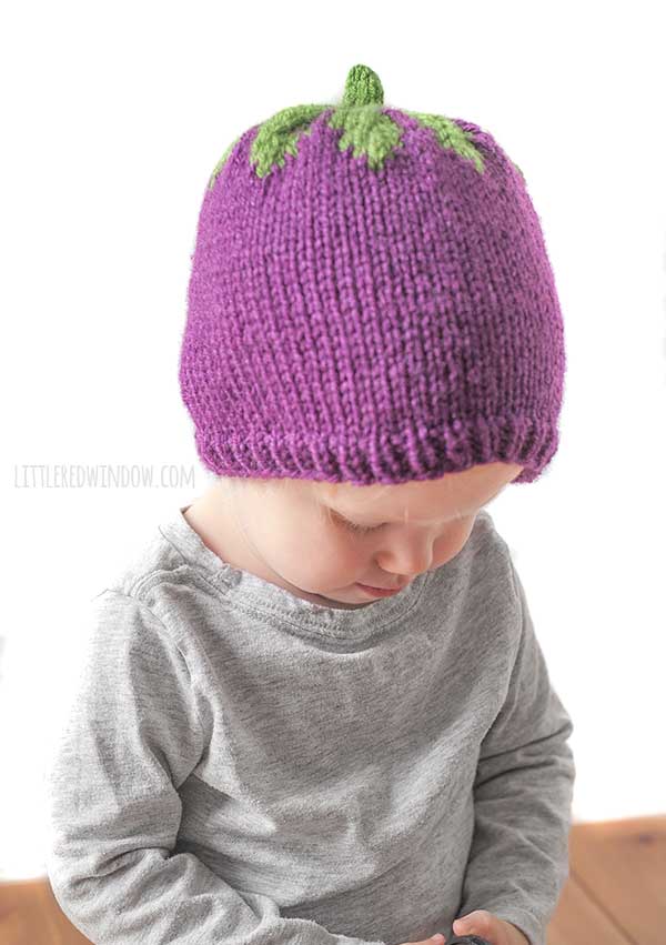 baby in gray shirt examining something in their hands and wearing a purple and green knit eggplant hat