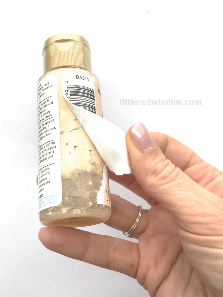 hand peeling back the label on a bottle of beige craft paint showing that the paint inside has separated