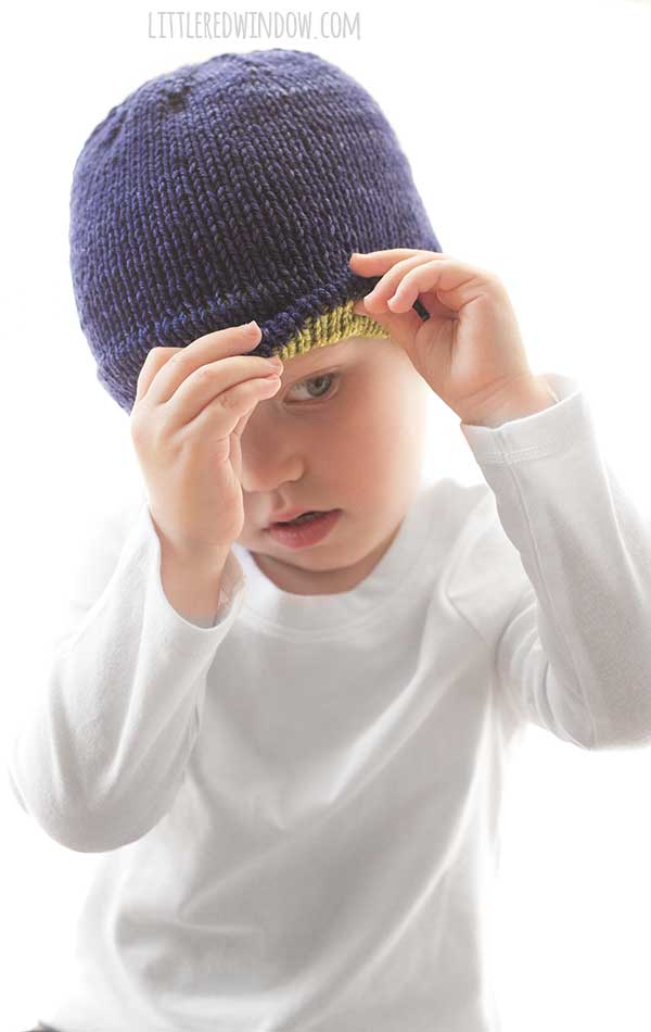 child wearing white shirt lifting up the brim of their navy blue knit hat to reveal lime green inside and trying to look at it