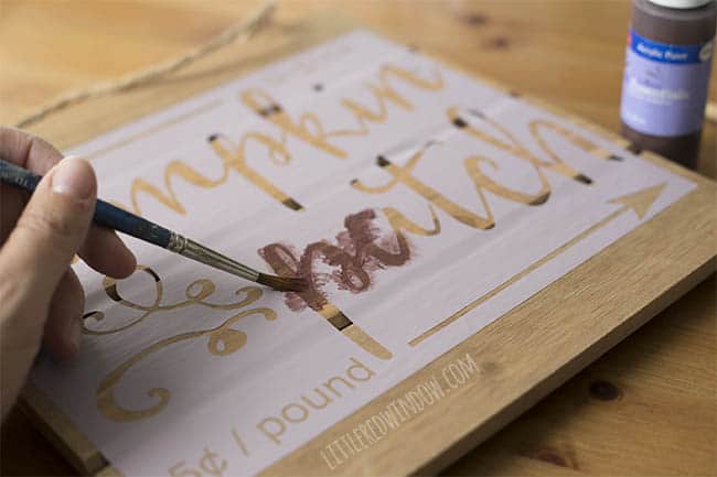 persons hand holding a paintbrush and painting the stencil on top of the wood with brown paing