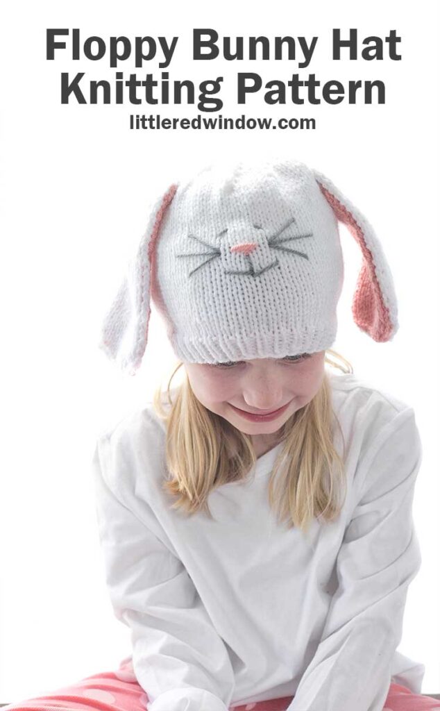 Smiling girl in white shirt wearing a white knit hat with long floppy white bunny ears with pink lining and a cute bunny face on the front looking down at her lap