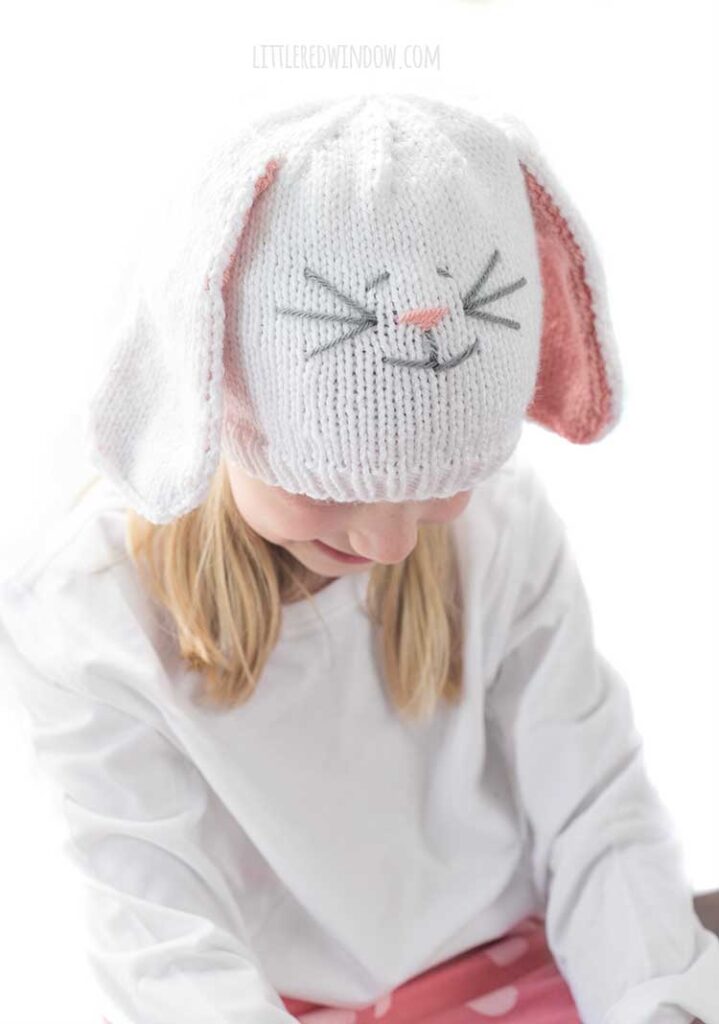 Smiling girl in white shirt wearing a white knit hat with long floppy white bunny ears with pink lining and a cute bunny face on the front looking off to the left