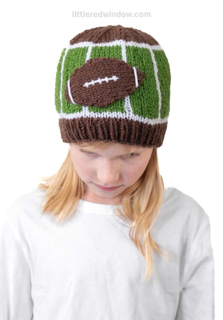 A child wearing a football hat styled like a knit football field, complete with a brown football decoration, stands against a white background and looks down.