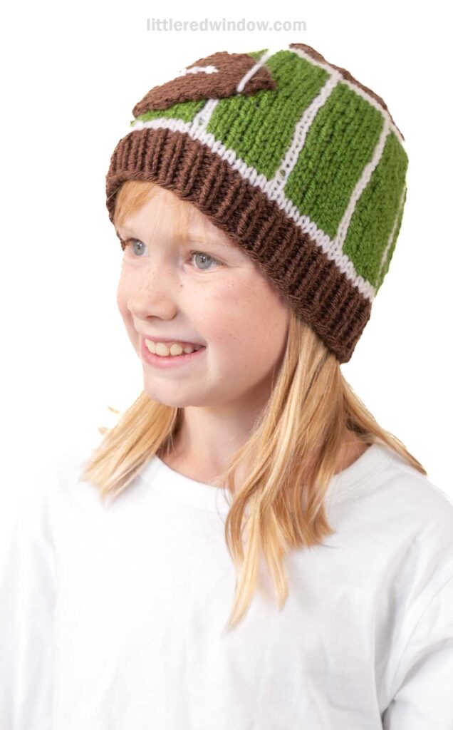 Smiling child with blonde hair wears a playful football hat resembling a field, paired with a plain white shirt against a white background.