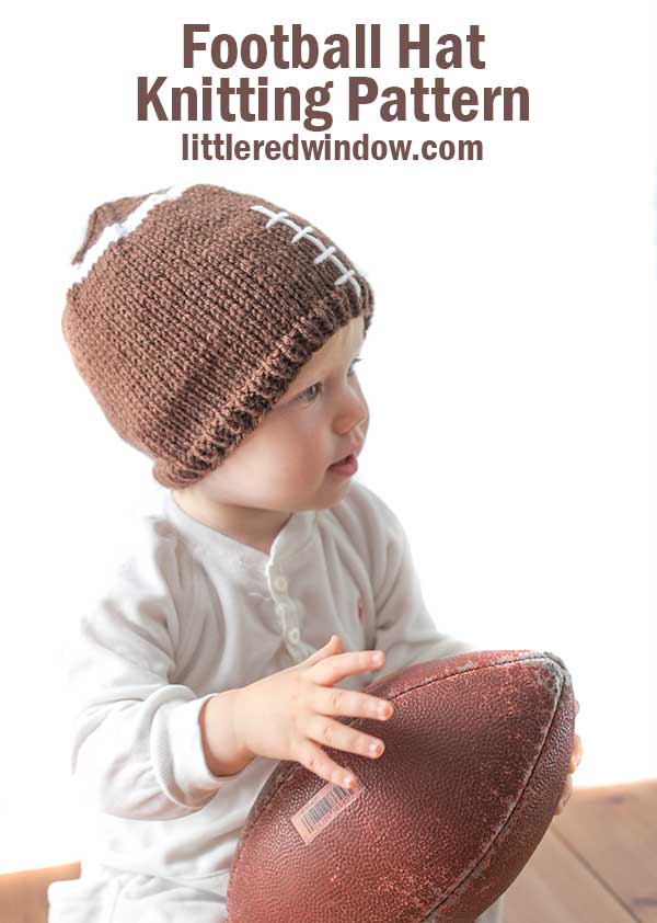 baby wearing brown knit hat that looks like a football and holding a real football in her lap looking off to the right