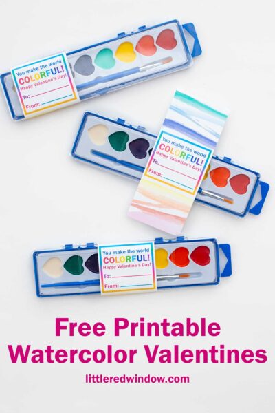 Three watercolor paint sets in blue cases are displayed. Each has a card labeled "You make the world COLOR-FULL. Happy Valentine's Day! To: From:" The background is white, and text below reads "Free Printable Watercolor Valentines" with a website link.