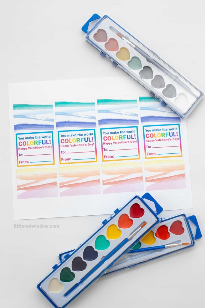 Three watercolor palettes with various colors are placed next to a printed sheet of Valentine's Day cards. The cards say