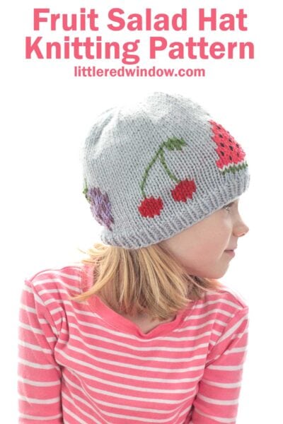 A child wears a light gray fruit salad hat with playful fruit designs and a pink-and-white striped shirt, with text above reading "Fruit Salad Hat Knitting Pattern" and "littleredwindow.com.