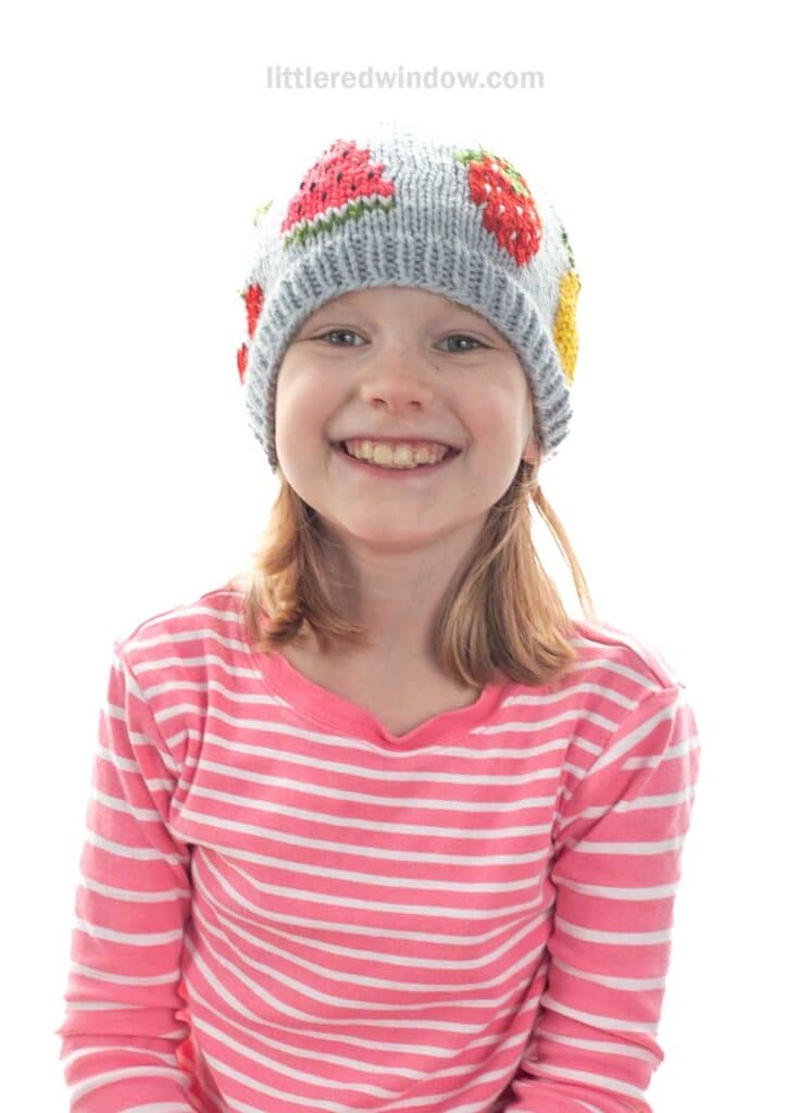 Smiling young girl wearing a pink and white striped shirt and a gray knit fruit salad hat decorated with colorful fruit designs, including strawberries. The background is plain white.