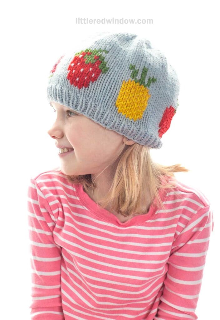 A young girl in a pink striped shirt smiles while wearing a light gray fruit salad hat decorated with colorful fruit designs, including strawberries and pineapples, against a white background.