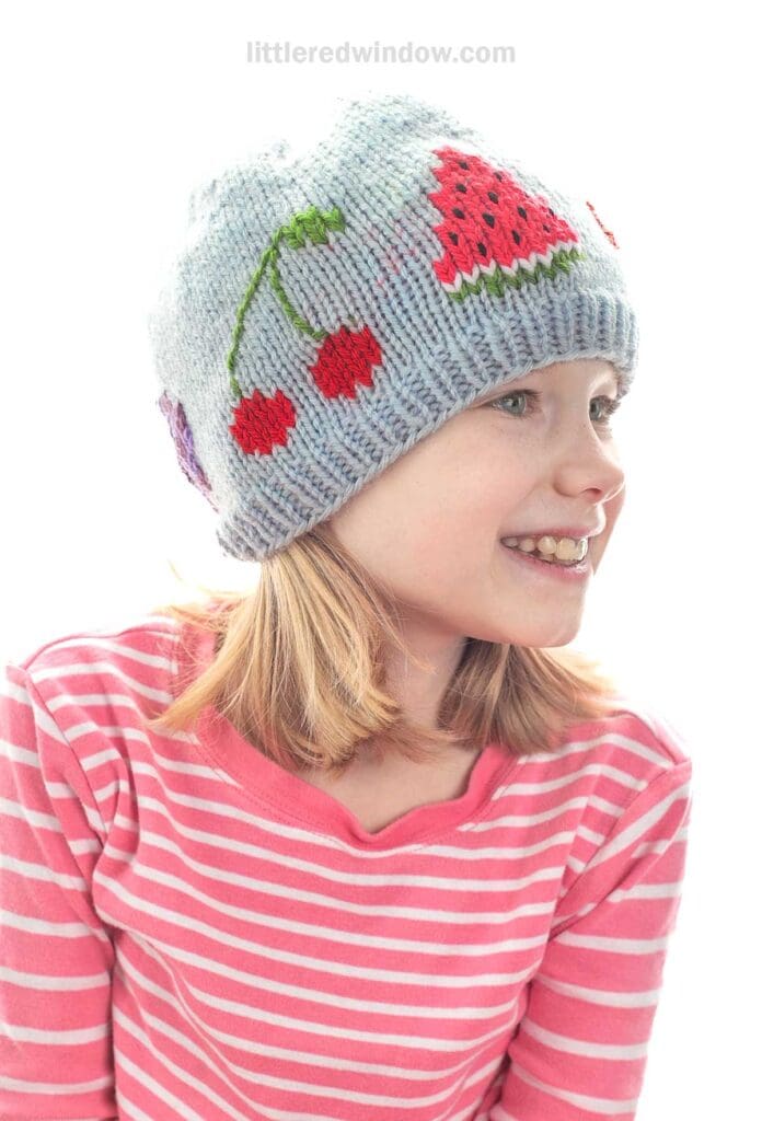 Smiling child in a pink and white striped shirt wears a light gray fruit salad hat decorated with colorful fruit designs, including cherries and a watermelon slice, on a white background.