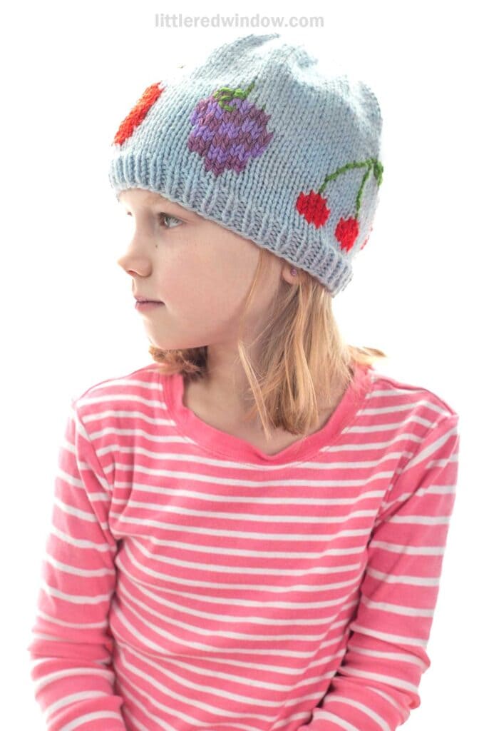 Young girl in a pink and white striped shirt wears a light blue knit fruit salad hat, decorated with colorful fruit designs like cherries, a strawberry, and a purple grape. She is looking to the left.
