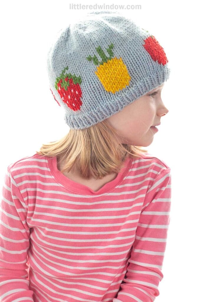 A young child wearing a gray fruit salad hat decorated with colorful fruit patterns and a pink striped shirt, looking to the side against a white background.