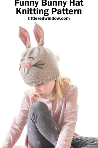 A child sports a gray knit hat with adorable bunny ears and a pink nose, perfectly capturing the essence of a rabbit. Above, the text says, "Funny Bunny Hat Knitting Pattern littleredwindow.com." Dressed in a pink top and gray pants, the child sits contentedly.
