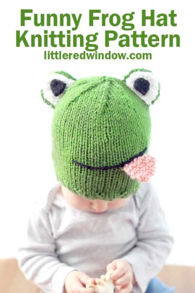 Baby holding giraffe toy in gray shirt wearing a green knit hat that looks like a frog with eyes on top and smiling mouth with pink tongue sticking out in front of a white background looking down