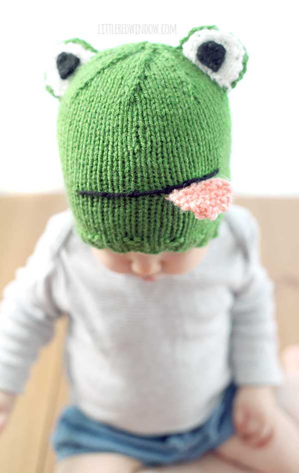 Baby holding giraffe toy in gray shirt wearing a green knit hat that looks like a frog with eyes on top and smiling mouth with pink tongue sticking out in front of a white background looking down
