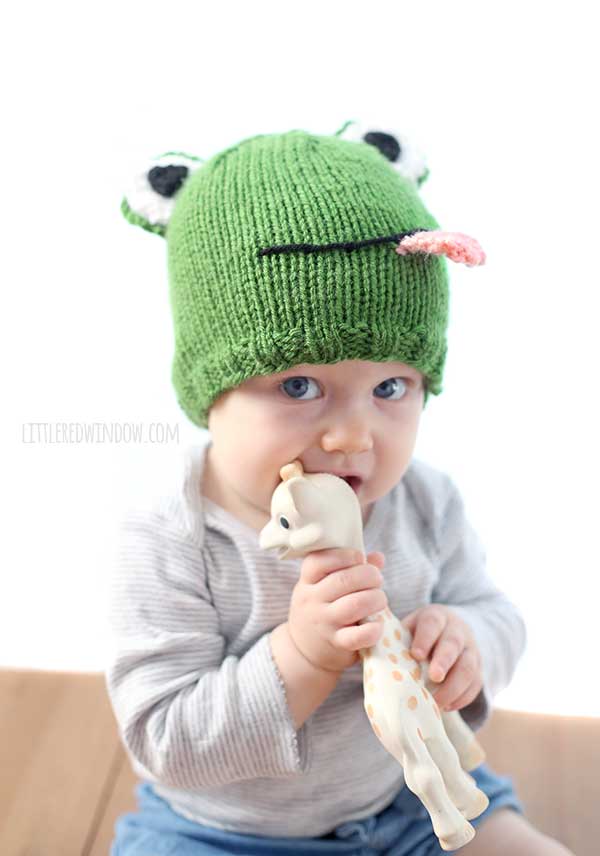 baby chewing on giraffe toy while wearing a green knit frog hat with eyes on top and a pink tongue