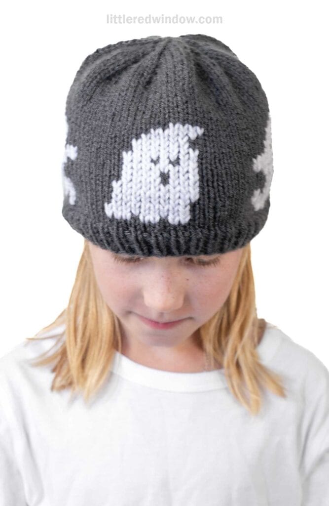 A child with blonde hair wearing a dark gray football hat featuring a white ghost pattern, looking down while dressed in a plain white shirt.
