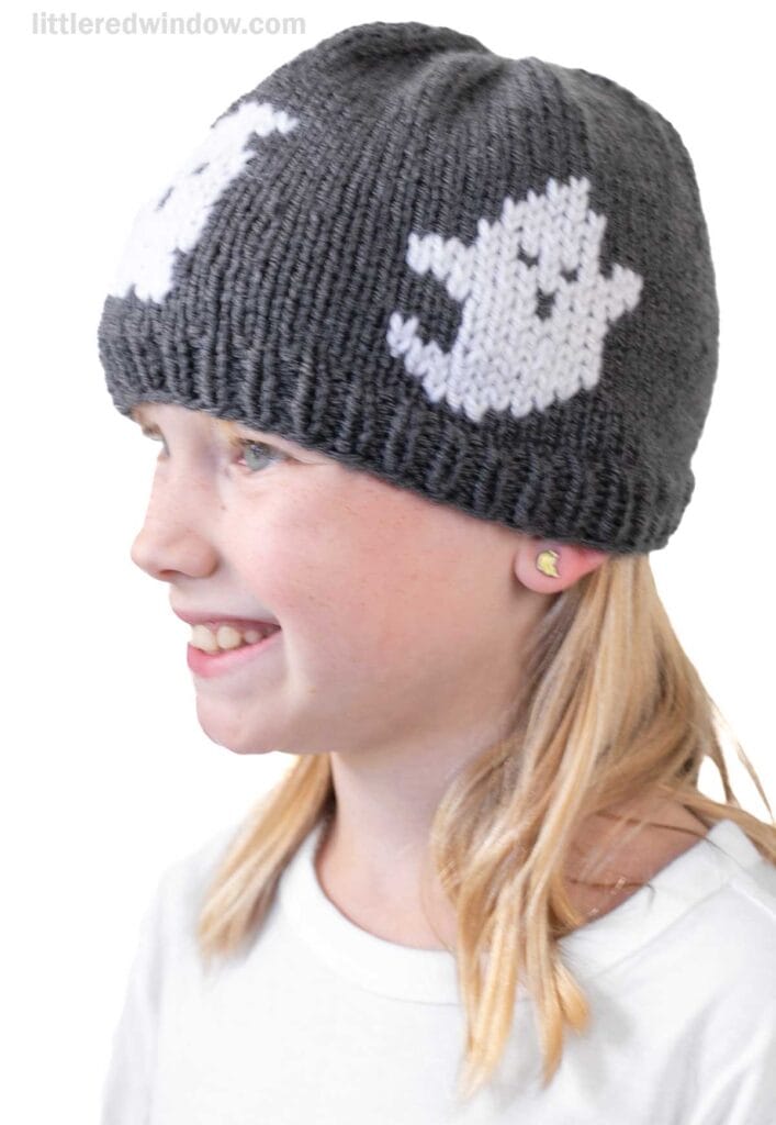 A young girl wearing a gray knit football hat with white ghost designs on it, smiling and facing left. She has blonde hair, small stud earrings, and is wearing a white shirt.