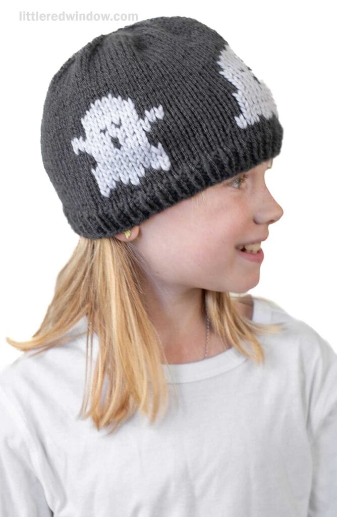A smiling child with blonde hair wears a dark knit football hat featuring white ghost designs and a plain white t-shirt, looking to the right.